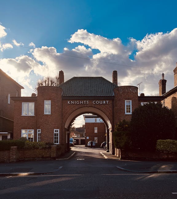 The photograph shows the entrance to Knights Court, a historic brick building with an arched gateway. The structure features two symmetrical square buildings on either side of the arch, each with multiple white-framed windows. The arch itself is topped with a slightly sloped, tiled roof and the words 'KNIGHTS COURT' are prominently displayed above the archway. In the foreground, a paved driveway leads through the arch, with a few parked cars visible within the court area. To the right and left of the entrance, there are well-maintained bushes and low brick walls. The background reveals a partly cloudy sky with patches of blue and soft sunlight casting shadows on the building facade. This setting, with its classic architecture and urban environment, is consistent with a location involved in home relocation or furniture transport services, as provided by companies like Man and a Van Knightsbridge, especially when preparing for a house removal or moving process.
