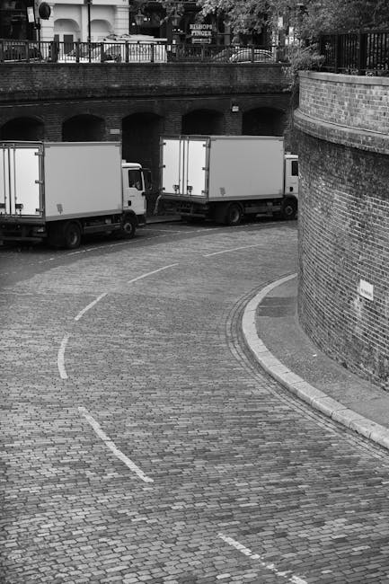 A street scene showing two delivery vans parked on a cobblestone pavement near an arched brick underpass, with a curved wall on the right side. The image captures a loading or unloading area potentially used for home relocation or furniture transport, with no visible personnel. The pavement has white dashed lines indicating parking or loading zones, and the background includes a railing and some trees, suggesting an urban environment suitable for house removal services. The scene illustrates the logistical aspect of a moving process, possibly coordinated by Man and a Van Knightsbridge, involving vehicle access for removals and transport of household items.