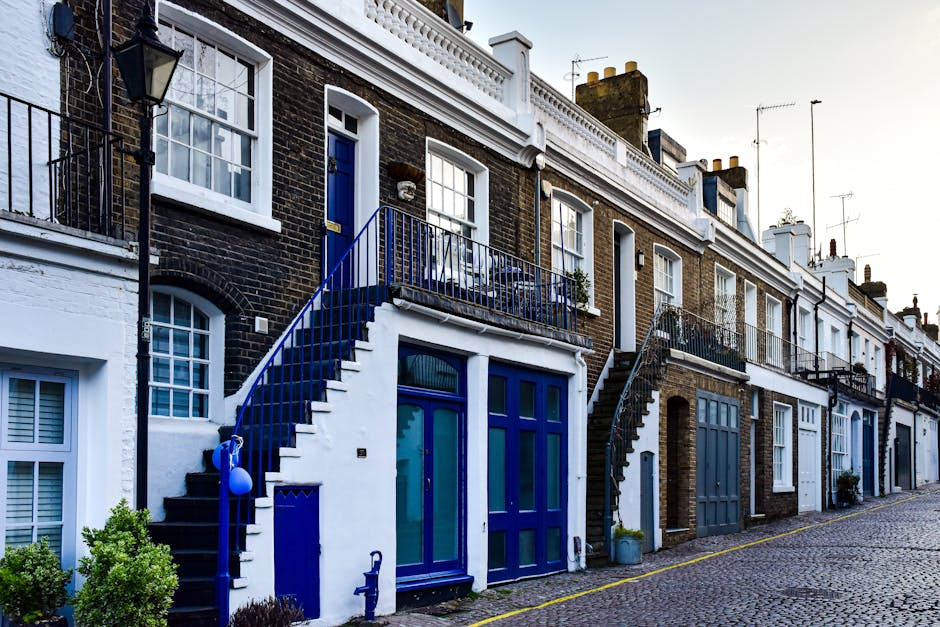 A row of terraced houses with brick facades and white window frames, situated along a cobbled street. The property in the foreground has a white-painted ground floor with large blue-framed glass doors and a small outdoor staircase with a blue metal railing leading up to a first-floor balcony. The balcony is also secured with a matching blue metal railing and decorated with a small plant. There are cardboard boxes, wrapped furniture, and packing materials visible on the pavement near the entrance, indicating a home relocation process. A van is partially visible on the street, adjacent to the curb, suggesting furniture transport and moving logistics activities. The scene captures the typical setting for house removals in a historic urban area, with ambient daylight illuminating the environment, which is part of a professional moving service performed by Man and a Van Knightsbridge.
