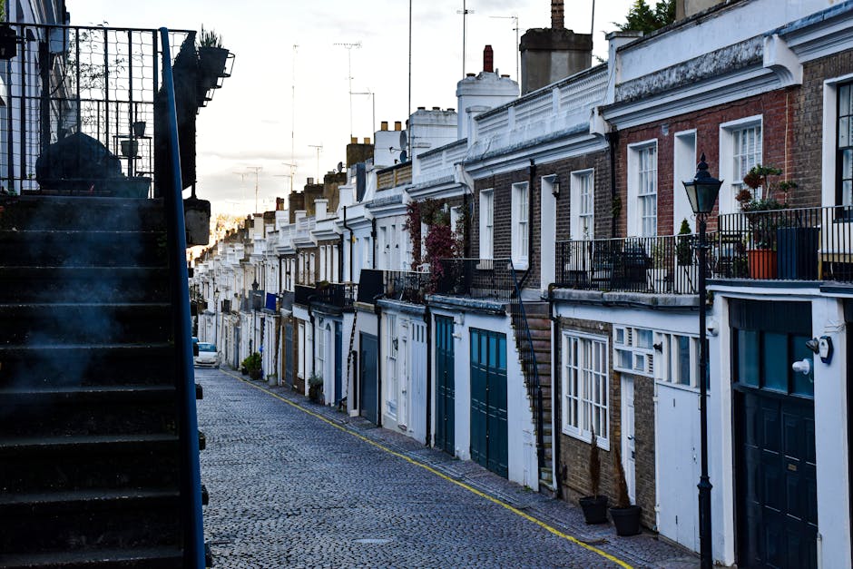 A row of terraced houses with brick facades and white window frames, situated along a cobbled street. The property in the foreground has a white-painted ground floor with large blue-framed glass doors and a small outdoor staircase with a blue metal railing leading up to a first-floor balcony. The balcony is also secured with a matching blue metal railing and decorated with a small plant. There are cardboard boxes, wrapped furniture, and packing materials visible on the pavement near the entrance, indicating a home relocation process. A van is partially visible on the street, adjacent to the curb, suggesting furniture transport and moving logistics activities. The scene captures the typical setting for house removals in a historic urban area, with ambient daylight illuminating the environment, which is part of a professional moving service performed by Man and a Van Knightsbridge.