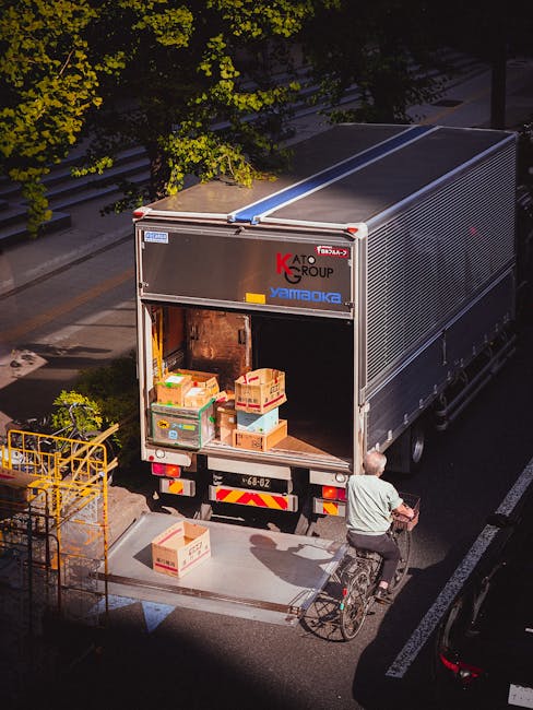A large moving truck with an open rear door, parked on a city street at dusk, loaded with cardboard boxes and plastic containers filled with household items ready for a home relocation. The truck is branded with the logos of Kato Group and Yamato, and positioned close to the pavement next to a bicycle rider who is about to load or unload items. Nearby, a yellow metal barricade is visible, suggesting access control around the loading area. The street is lined with trees, with some green foliage visible in the background, and the surrounding environment appears to be part of an urban residential area. The scene captures the logistics of furniture transport and packing during a professional removals service, with natural lighting emphasizing the process of loading or unloading in a residential setting, as facilitated by Man and a Van Knightsbridge.