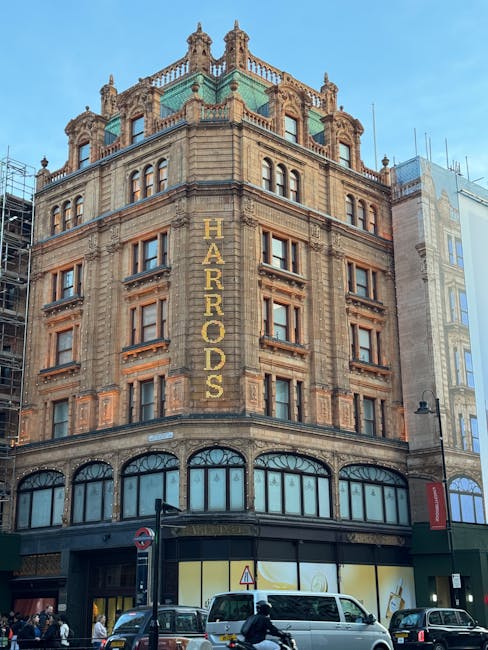 A tall, historic building located on a busy city street, showcasing ornate stonework and large arched windows at ground level. The building's facade features carved decorative elements and multiple floors with rectangular windows framed by detailed masonry. A prominent vertical yellow sign reading 'HARRORDS' is affixed to the upper part of the building, indicating a commercial establishment. Several vehicles, including cars and a van, are parked or moving along the pavement in front of the building, while pedestrians walk nearby. The street scene highlights typical urban activity surrounding a prominent structure involved in house removals and relocation services. The building is part of the logistics environment addressed in the Knightsbridge house removals guide by Man and a Van Knightsbridge.