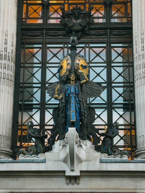 A detailed view of a decorative building façade featuring an ornate central sculpture with a winged figure in a blue dress, surrounded by smaller sculptures of seated figures and animal motifs, set against a background of large glass panels framed by classical columns. The sculpture is positioned above an entrance area, with architectural elements such as carved stone and metalwork visible. Though not directly related to house removals or furniture transport, this image illustrates architectural features that may be encountered during property relocations in London, such as Knightsbridge, where Man and a Van Knightsbridge offers professional local removal services.