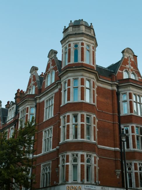 A multi-storey red brick building with ornate architectural features, including tall, narrow windows with white framing, decorative stonework, and a central cylindrical turret topped with a balcony and balustrade. The building is situated on a street corner with a black streetlamp nearby and a tree partially visible in the lower left corner. The sky above is clear and light blue. The scene depicts an urban residential area, consistent with historic or period properties often involved in house removals or property relocations, as managed by companies like Man and a Van Knightsbridge, whose services may include packing, furniture transport, and loading processes within such buildings.