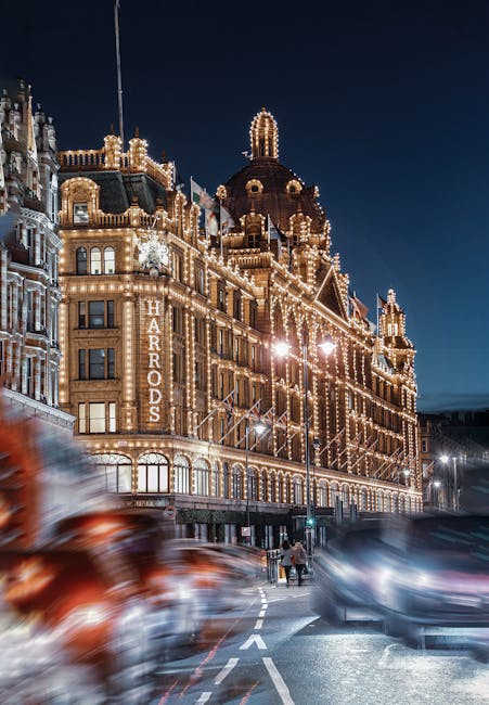 A nighttime view of the Harrods building in Knightsbridge, London, illuminated with numerous decorative and functional lights outlining its historic architecture, including ornate facades and domed towers. In the foreground, several blurred vehicles move along the street, indicating motion and bustling activity, with a few pedestrians crossing at a nearby crosswalk. The scene captures the vibrant city environment and the iconic presence of Harrods, which is relevant to house removals, packing, and moving services. The image’s lighting highlights the detailed exterior of the building, while the slightly blurred street activity emphasizes the busy urban context where professional removals, such as those by Man and a Van Knightsbridge, facilitate home relocation and furniture transport projects.