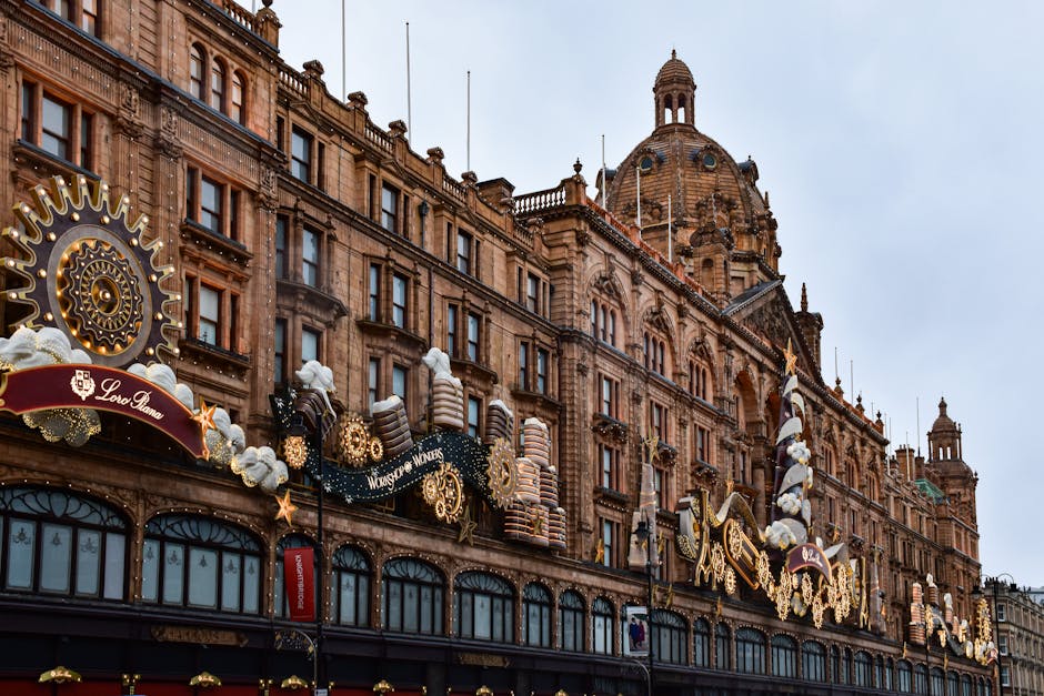 A nighttime view of the Harrods building in Knightsbridge, London, illuminated with numerous decorative and functional lights outlining its historic architecture, including ornate facades and domed towers. In the foreground, several blurred vehicles move along the street, indicating motion and bustling activity, with a few pedestrians crossing at a nearby crosswalk. The scene captures the vibrant city environment and the iconic presence of Harrods, which is relevant to house removals, packing, and moving services. The image’s lighting highlights the detailed exterior of the building, while the slightly blurred street activity emphasizes the busy urban context where professional removals, such as those by Man and a Van Knightsbridge, facilitate home relocation and furniture transport projects.