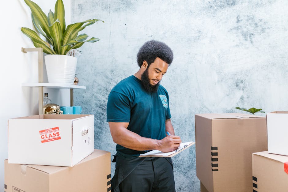 A professional removalist from Man and a Van Knightsbridge is indoors, standing between a collection of packed cardboard boxes and a potted green plant on a white shelf. The move organiser is wearing a navy blue t-shirt and black work pants, holding a clipboard and writing notes during a home relocation planning process. The background features a textured, light-colored concrete wall, illuminated by natural light, indicating an organized packing and preparing stage for furniture transport and loading for house removals. The scene captures the meticulous coordination involved in packing and moving services, with the boxes carefully sealed with packing tape, some labelled with contents, in a setting suited for a comprehensive house or office move.