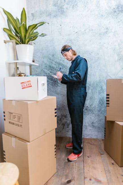 A man wearing a white t-shirt and a black baseball cap is standing inside a storage area or loading bay, writing notes or a checklist on a clipboard with a pen. Behind him, stacked cardboard moving boxes of various sizes are visible, some sealed with packing tape. The boxes are arranged on the floor and against the wall, indicating preparation for a house or office relocation. In the background, part of a reflective window or door is visible, providing natural light that illuminates the scene. This setting illustrates the packing and loading process involved in professional removals services, such as those offered by Man and a Van Knightsbridge, supporting efficient furniture transport and home or office moves.
