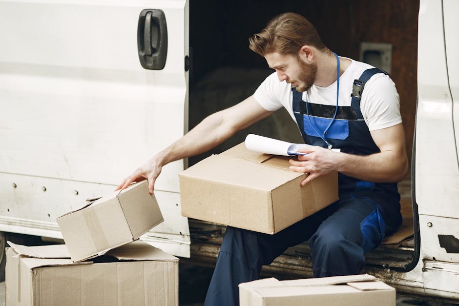 A male removal worker with light brown hair and a beard, dressed in a white t-shirt and navy overalls with blue accents, is seated at the open side door of a white delivery van during a home relocation process. He is carefully placing a cardboard box onto a pile of similar boxes, some on the ground and some inside the van. The worker holds a rolled-up document or packing list in one hand, indicating logistical or packing details related to the move. The van's interior is visible, with additional boxes and packing materials such as bubble wrap and plastic wrapping nearby, suggesting careful packing and transport of household items. The scene is set outdoors, with natural light illuminating the workspace, capturing the logistical details involved in furniture transport and packing during a professional removals service, as provided by Man and a Van Knightsbridge.