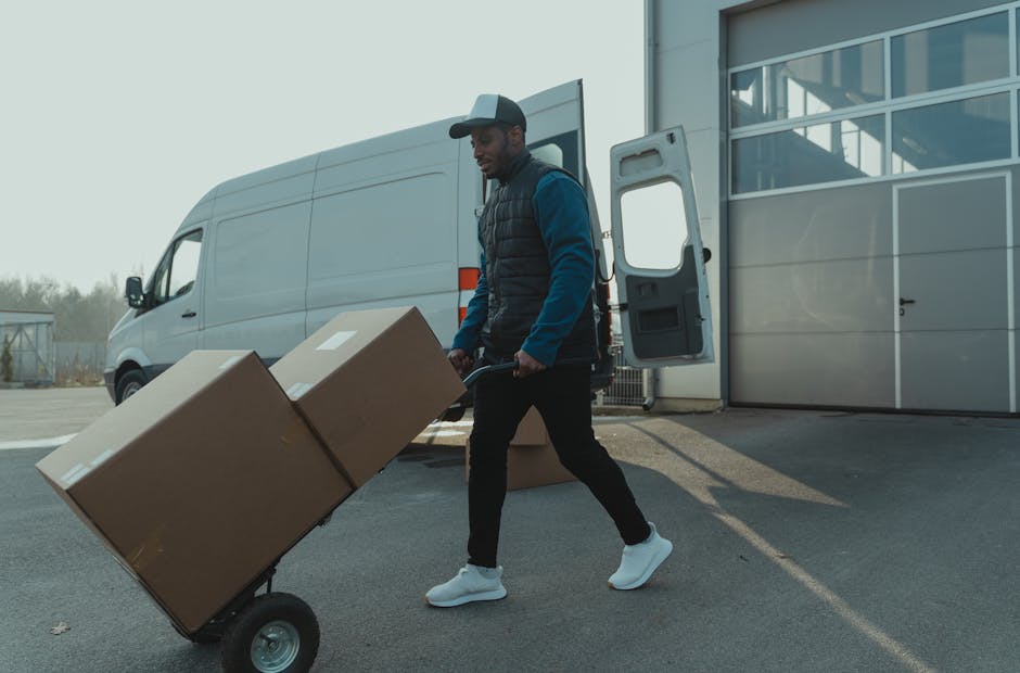 A man wearing a baseball cap, blue jacket, and black trousers is using a hand truck to transport two large cardboard boxes outdoors on a paved area next to a grey commercial van with its rear doors open. Inside the van, visible loading equipment and additional packaging materials are seen. The background includes a modern building with glass windows and a clear sky, indicating daytime. This scene illustrates the process of packing and loading household items for home relocation, as part of furniture transport and moving logistics handled by Man and a Van Knightsbridge, a professional removals service operating in the Knightsbridge area.