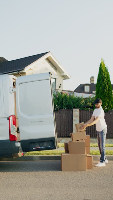 A male removal worker with light brown hair and a beard, dressed in a white t-shirt and navy overalls with blue accents, is seated at the open side door of a white delivery van during a home relocation process. He is carefully placing a cardboard box onto a pile of similar boxes, some on the ground and some inside the van. The worker holds a rolled-up document or packing list in one hand, indicating logistical or packing details related to the move. The van's interior is visible, with additional boxes and packing materials such as bubble wrap and plastic wrapping nearby, suggesting careful packing and transport of household items. The scene is set outdoors, with natural light illuminating the workspace, capturing the logistical details involved in furniture transport and packing during a professional removals service, as provided by Man and a Van Knightsbridge.