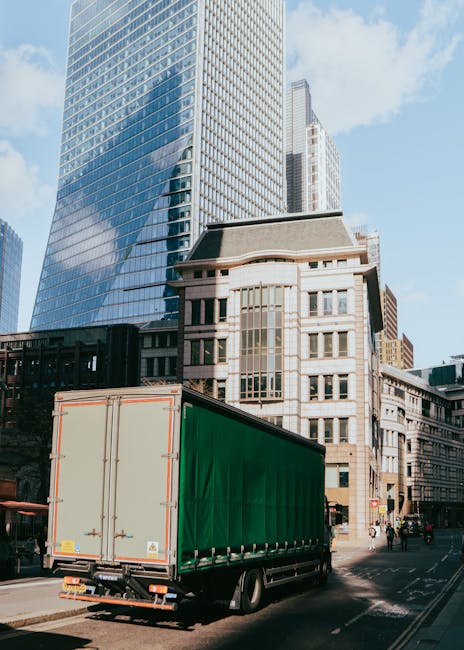 A large green curtain-sided delivery truck parked on a city street in Knightsbridge, London, with tall modern glass skyscrapers and historic stone buildings in the background. The truck is aligned parallel to the pavement near a busy area used for home relocation and furniture transport services. No loading activity or personnel are visible in the image. The scene is illuminated by natural daylight, highlighting the urban environment where professional removals, such as those by Man and a Van Knightsbridge, may occur during packing and moving processes within the city’s commercial district.